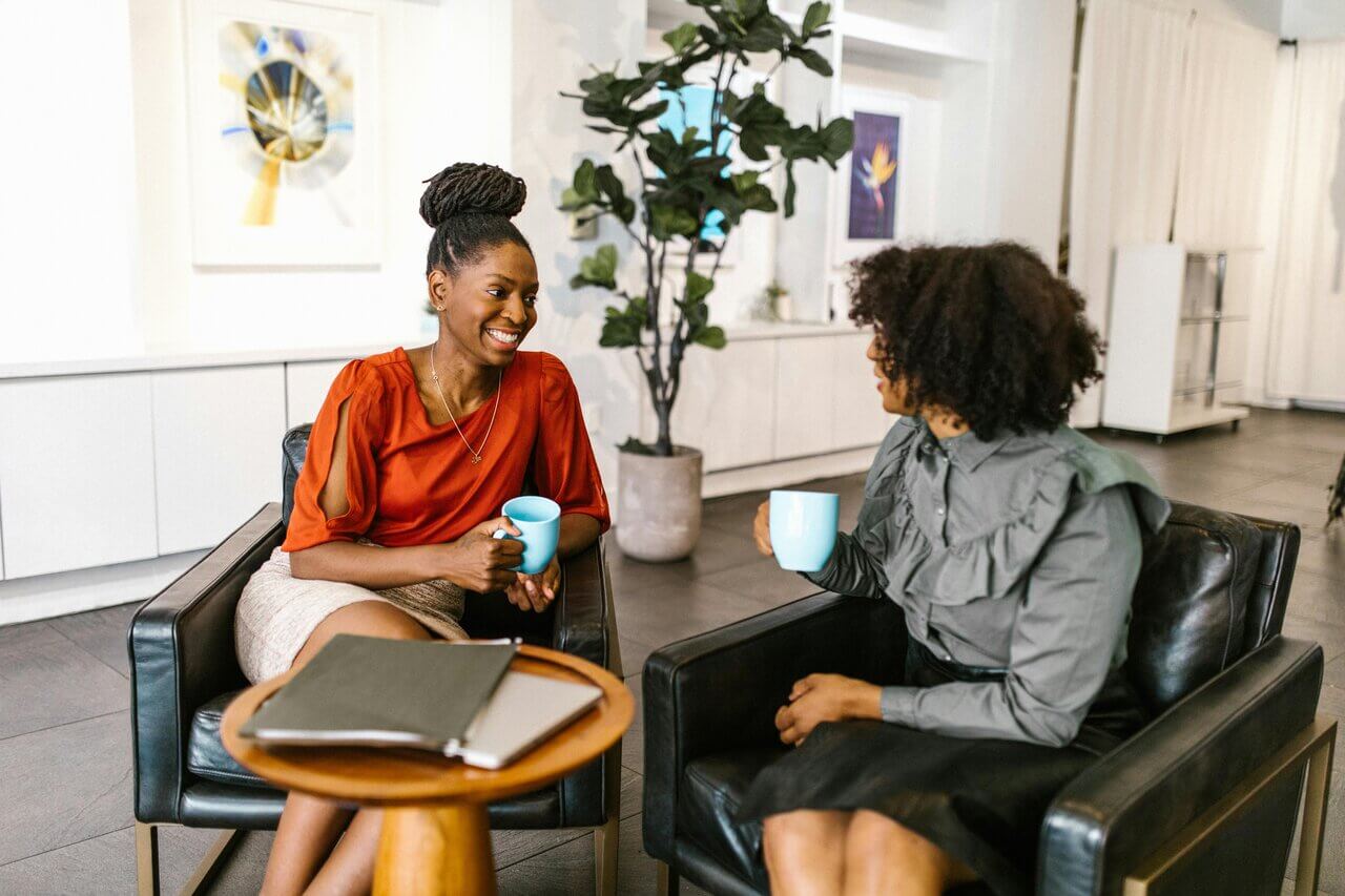 Two women in office sitting on armchair with coffee cup conversating